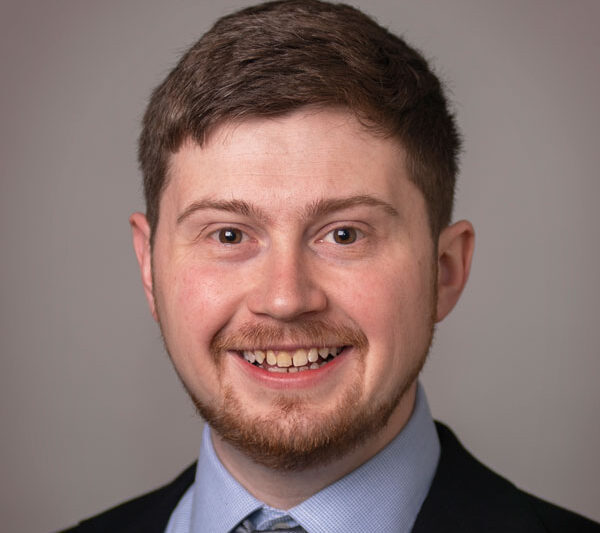 Alexander Dowling, smiling in a professional headshot. He has short brown hair, a trimmed beard, and is wearing a dark suit jacket, light blue dress shirt, and gray tie. Neutral background.