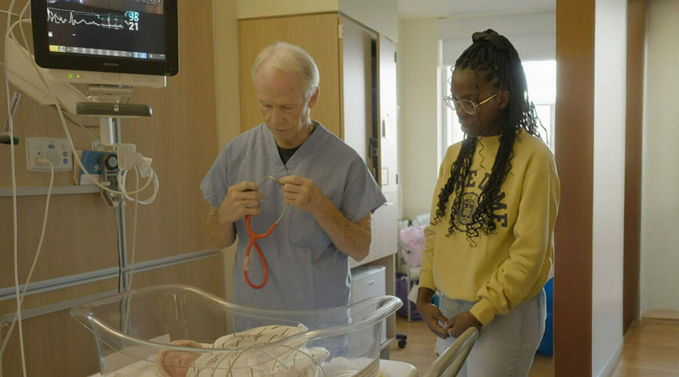 A doctor in blue scrubs holds a red stethoscope, looking down at a newborn lying in a hospital bassinet. A young woman in a yellow Notre Dame sweatshirt stands next to the bassinet.