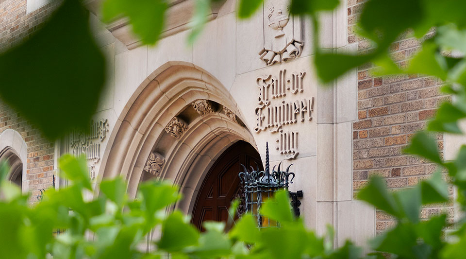 Ornate stone entrance to Cushing Hall of Engineering, with the building name engraved in gothic-style lettering above a large arched wooden door, partially framed by green leaves in the foreground.