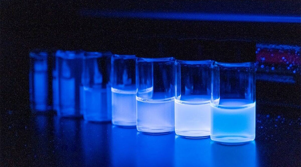 Several glass vials containing bright blue fluorescent liquid on a dark surface under blue light.