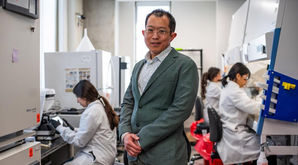 A smiling engineer in a green blazer and glasses stands in a lab with students in white coats working at equipment.