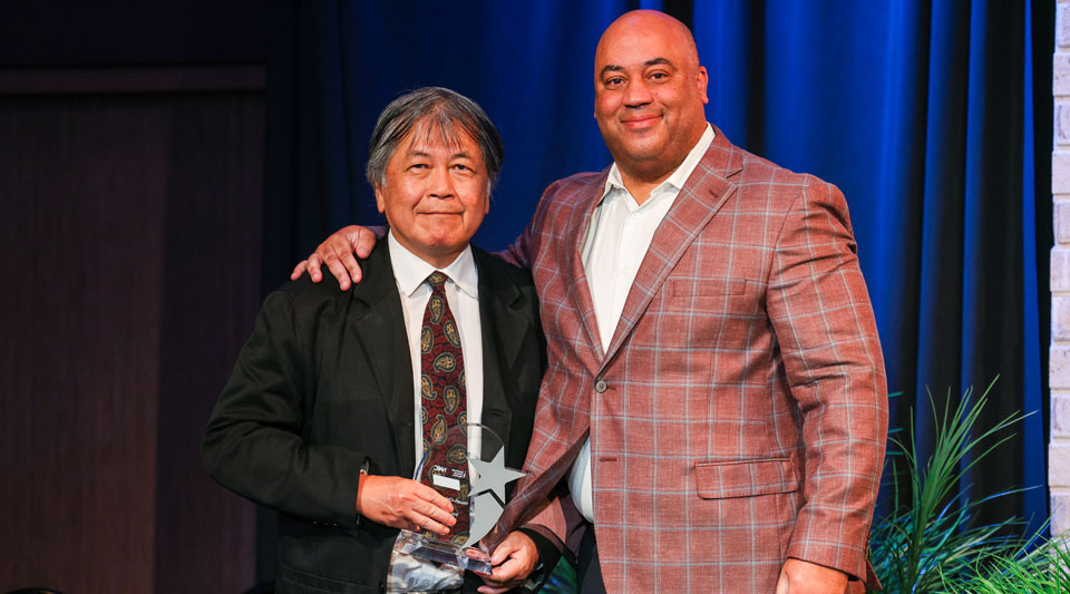 Chia Chang stands smiling and holding an award. Next to him is Notre Dame head men’s basketball coach, Micah Shrewsberry. Micah's arm is around Chia's shoulders.