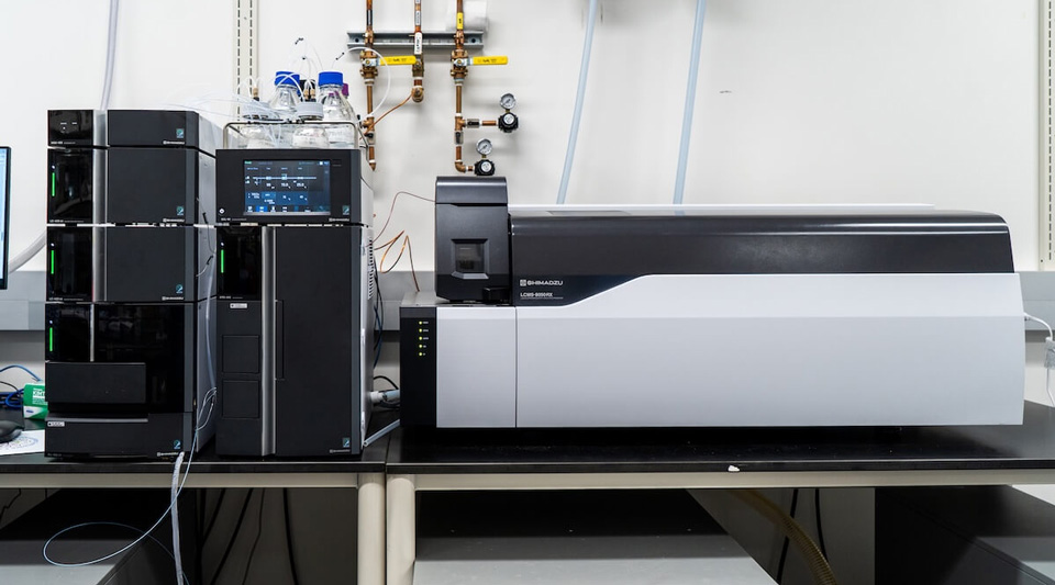Black Shimadzu LC modules with bottles, white and black LCMS-8050RX mass spectrometer, copper piping in a lab.